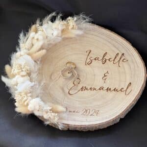 Image d'un rondin de bois clair servant de support pour des alliances. Les alliances sont posées sur un coussin en dentelle, entourées de décorations florales séchées dans les tons beiges et blancs. Le rondin est gravé des noms 'Isabelle & Emmanuel' et de la date 'mai 2024