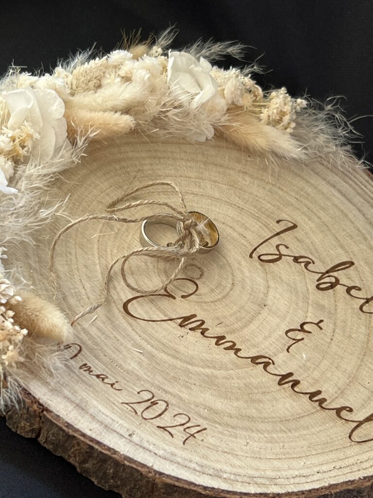 Image d'un rondin de bois clair servant de support pour des alliances. Les alliances sont posées sur un coussin en dentelle, entourées de décorations florales séchées dans les tons beiges et blancs. Le rondin est gravé des noms 'Isabelle & Emmanuel' et de la date 'mai 2024