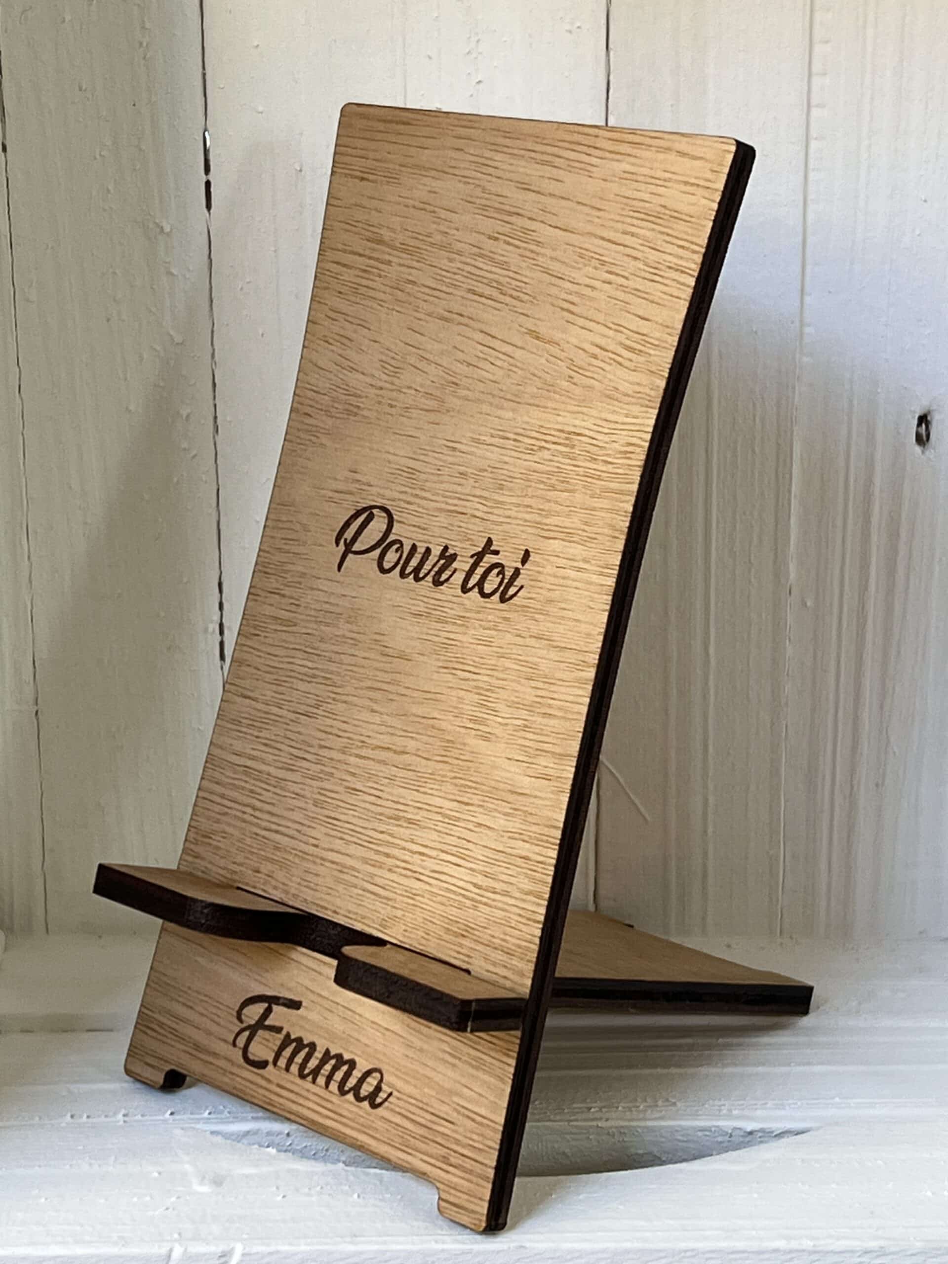 Image d'un support de téléphone en bois clair, avec un design simple et fonctionnel. Le support est gravé des mots 'Pour toi' sur la partie supérieure et 'Emma' sur la base. Le fond est clair et légèrement texturé